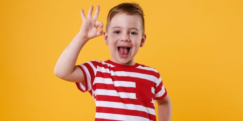 Portrait of an excited little boy standing and showing okay gesture isolated over orange background